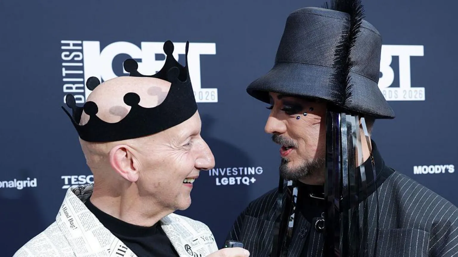A photo of Stephen Jones and Boy George from the shoulders up. Jones stands on the left, he has a bald head and is wearing a black crown made from a felt-style fabric. His jacket has a newspaper print and he wears a black top underneath. Boy George is wearing a pinstripe jacket and a black bucket-style hat with tassles. He has facial hair and gems around his eyes.