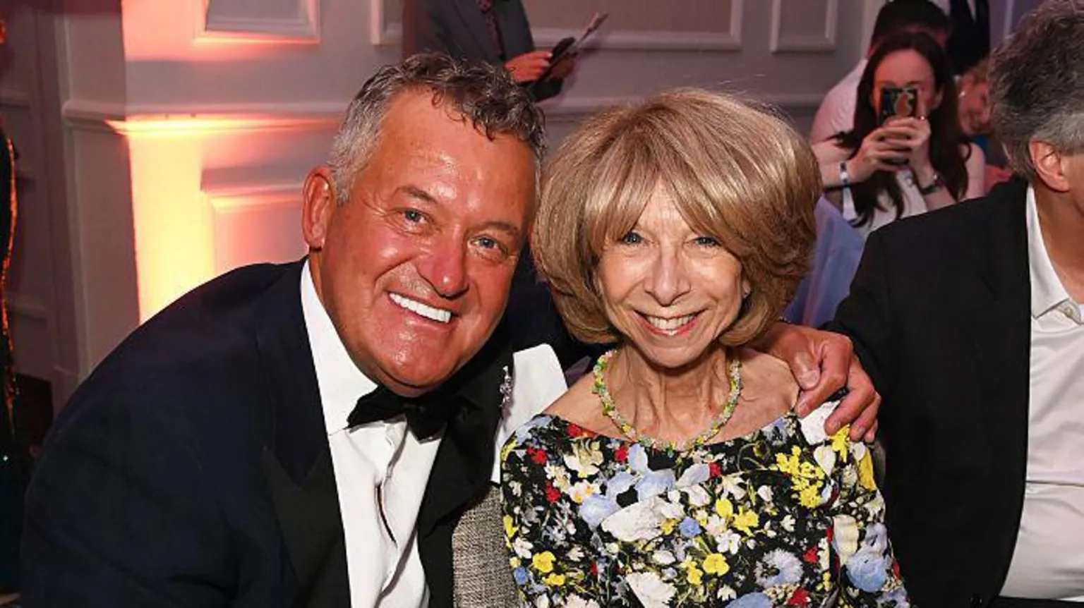 Paul Burrell, wearing a black suit jacket, white shirt and a black bow tie, poses with his arm around Helen Worth. She has short blond hair and is wearing a floral dress. Both are smiling.
