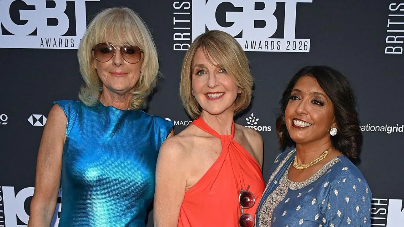 A photograph of Jane Moore, Kaye Adams and Sunetra Sarker from the waist up on the red carpet. The background is black with a logo reading 'British LGBT Awards'. Jane, on the left, wears a shiny blue dress and sunglasses with her hair in a blond bob. Kaye wears a red halter-neck dress with a similar hairstyle. Sunetra is on the right and wears a blue dress with gems, a gold necklace and silver earrings. She has dark hair.