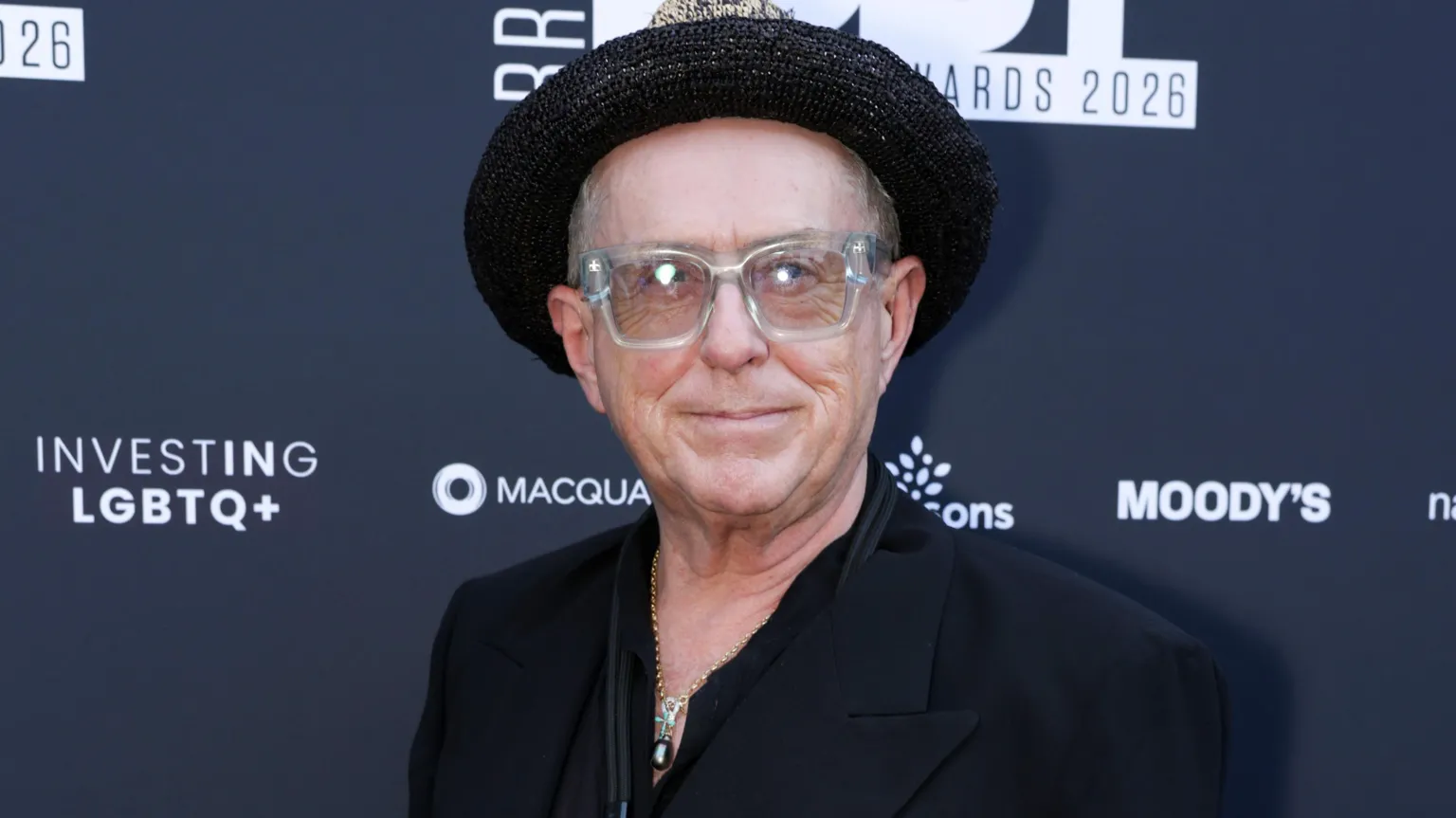 Holly Johnson on the red carpet, pictured here from the shoulders up. He is wearing a dark jacket, a chain around his neck, glasses with clear frames, and a hat.