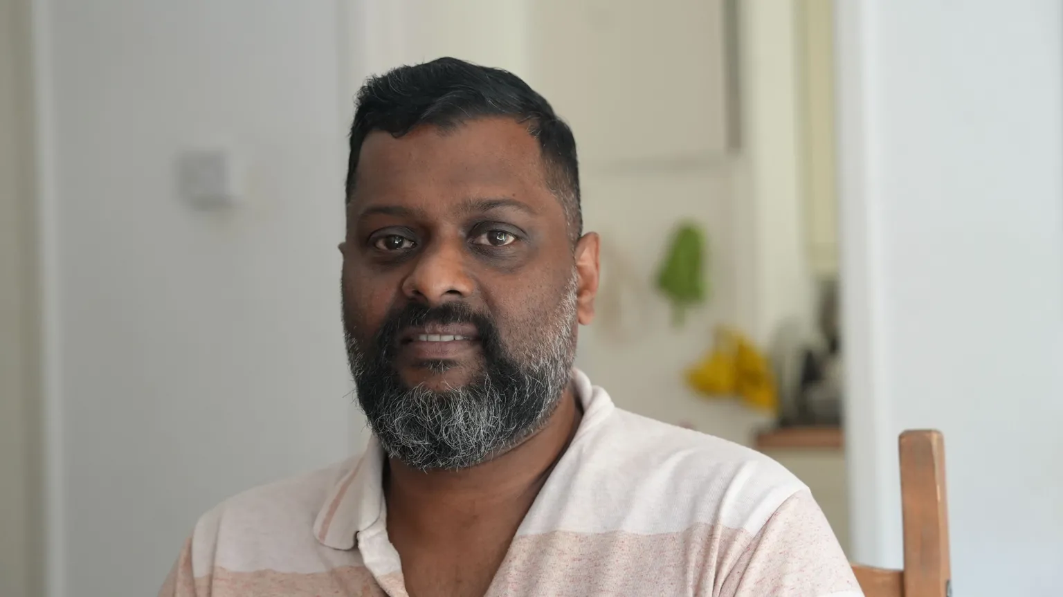 Sumesh has a dark beard and is wearing a beige polo shirt. He is sitting in a wooden chair and smiling at the camera