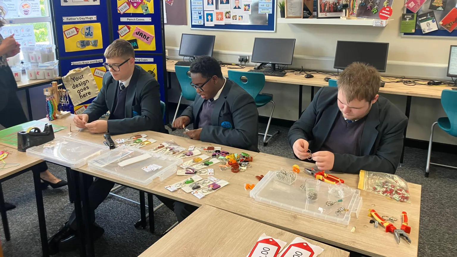 Pupils are sitting behind desks working with tools. There are keyrings on the desks. The pupils are wearing their school uniforms. Computers are behind them.