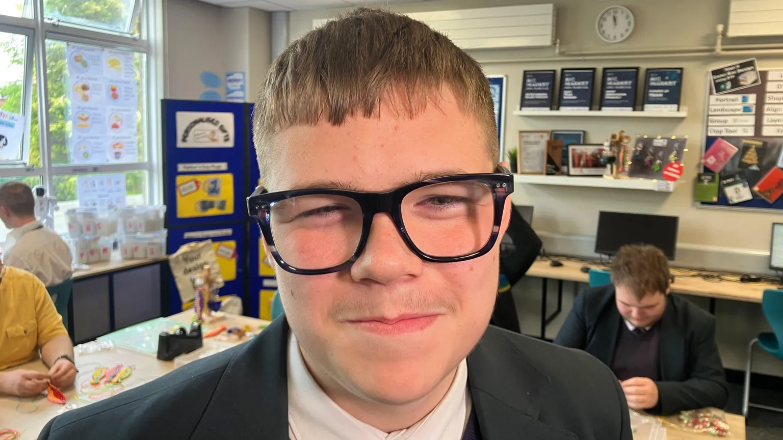 Darren has dark hair and wears glasses. He's wearing his school blazer with a white shirt. Pupils are sitting in the background behind desks. Displays are on walls.