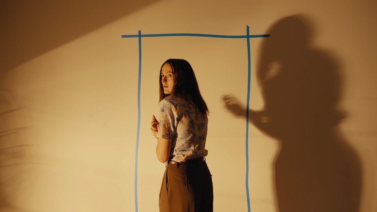 woman stands to the side by a wall, a shadowy silhouette reflected