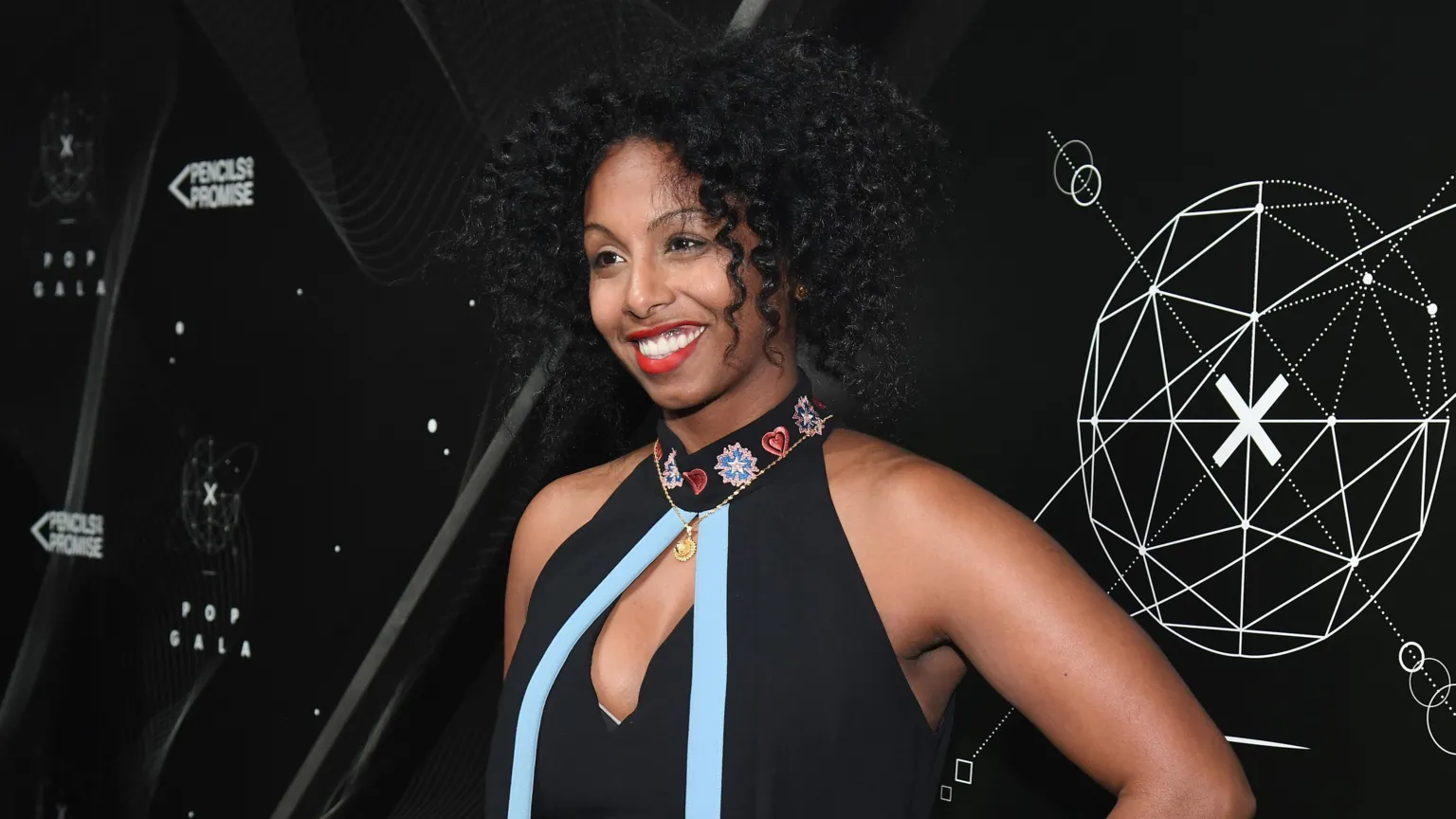 Luam wears a black halter neck dress with a blue ribbon on the front and flowers on the collar. She wears red lipstick and has her black curly hair in a short style