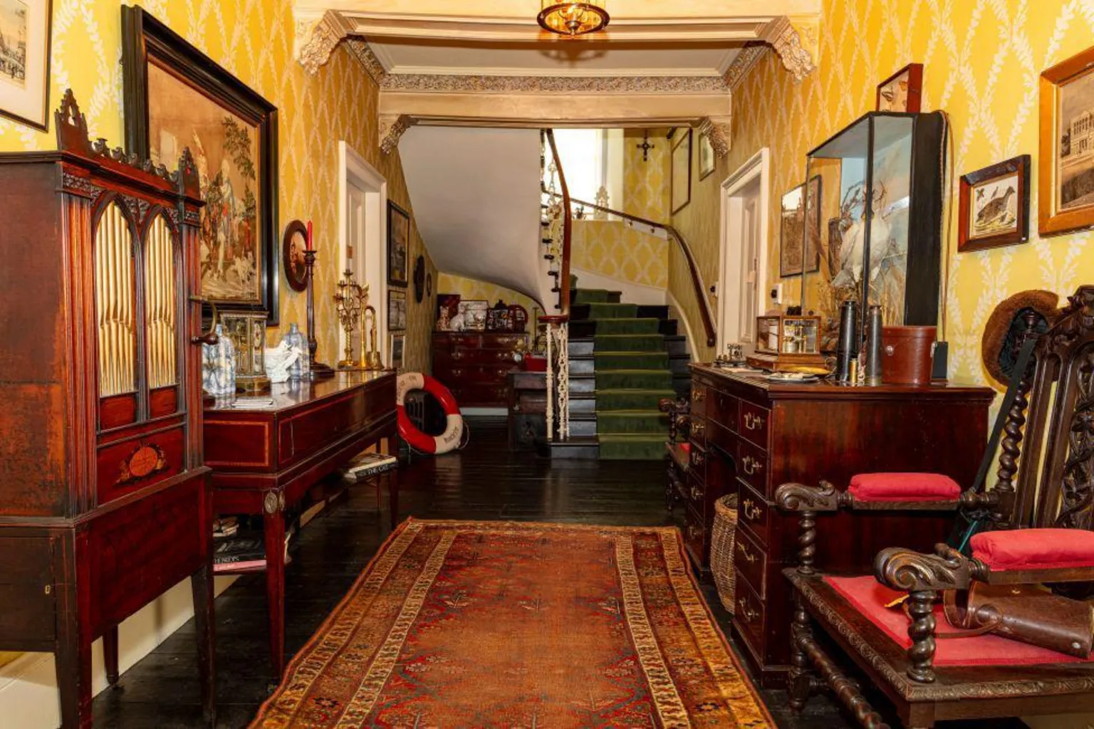 Steven Gourlay An old fashioned-looking interior of a building, featuring a winding staircase, chairs, sideboards, and an old rug on the floor.