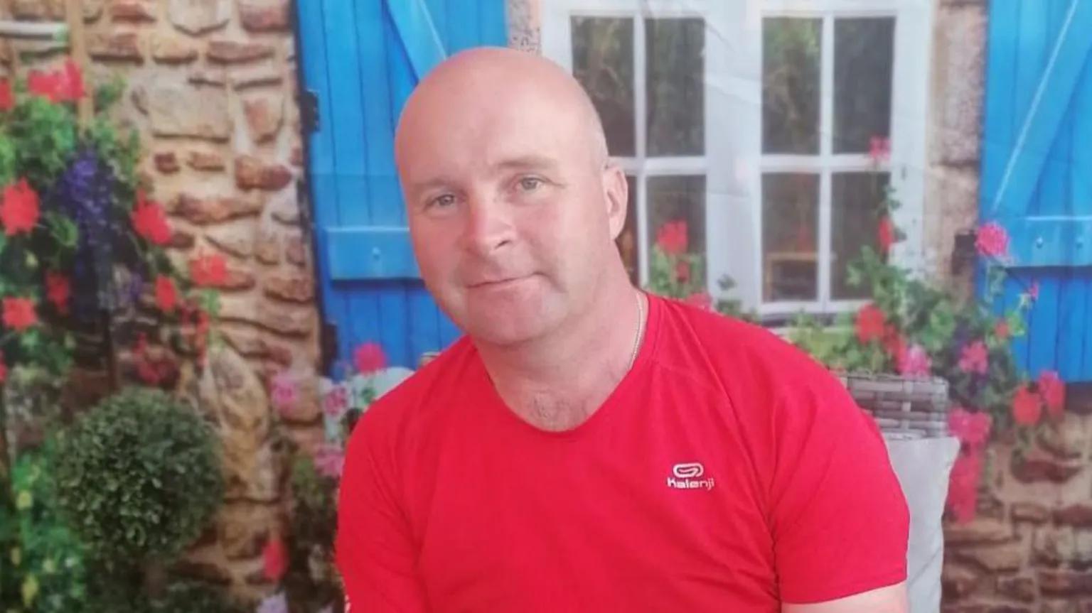 JP McDaid a man wearing a red t-shirt sitting in front of a house with blue shutters and flowers on the wall