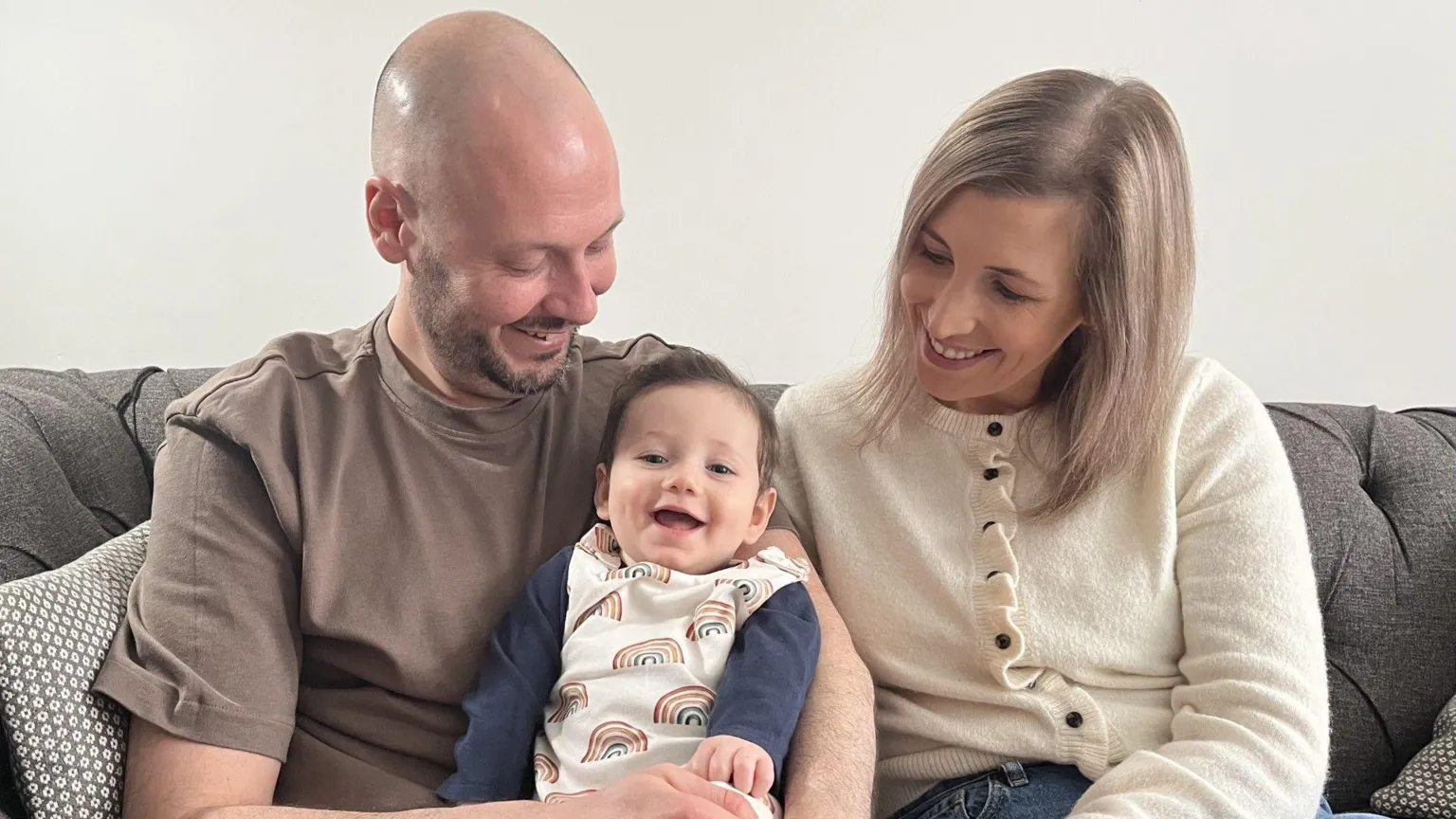 A bald man wearing a brown t-shirt and blue jeans sits next to a blonde woman wearing a cream cardigan and blue jeans. They are seated on a grey sofa and smiling down at a beaming baby who has dark hair and wears a blue top and rainbow-patterned romper suit.
