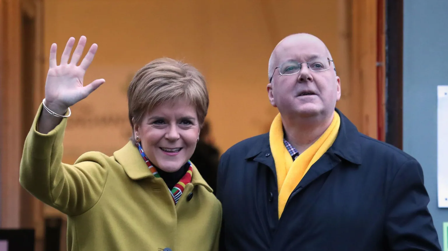  Nicola Sturgeon and Peter Murrell stand outside the entrance to a building marked with a sign reading “Polling Place Way In,” with a smaller sign below saying “Push bar to open.” Sturgeon, wearing a light green coat and colourful scarf, raises a hand in a wave, while Murrell wears a dark coat and bright yellow scarf and looks ahead. The open doorway behind them reveals warm indoor lighting, indicating they are just outside or entering a polling station.