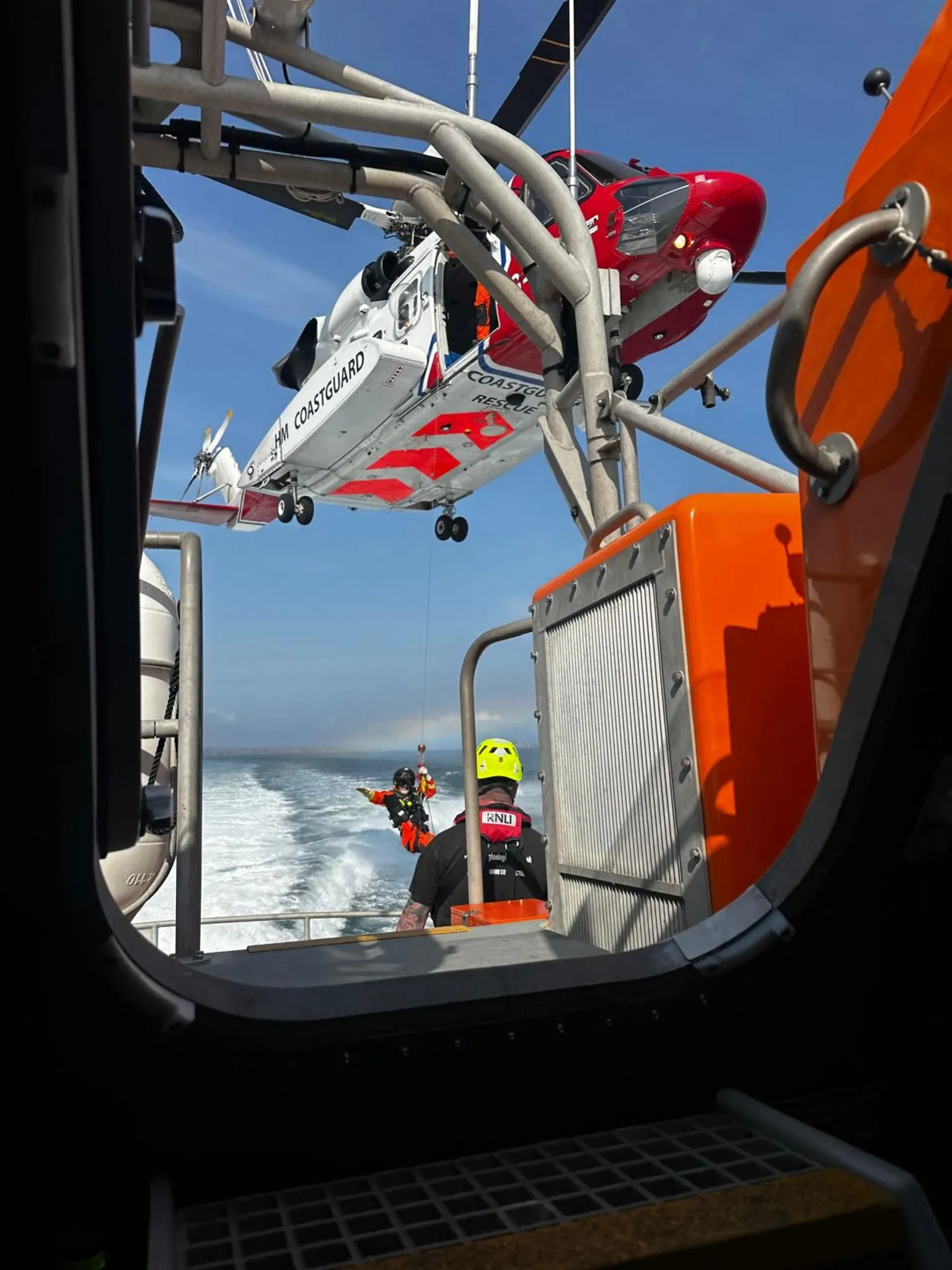 RNLI The paramedic wearing an orange suit and safety harness being lowered on to the lifeboat. The white and red helicopter is lowering the paramedic on to the boat as a RNLI volunteer watches on. 