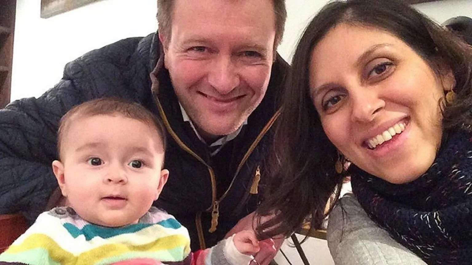 Family photo A selfie image of a man, a woman and a baby. The man is wearing a navy jacket and brown hair. A woman has dark hair and is wearing a grey jumper. The baby is wearing a multicoloured striped top and a little bit of dark hair.