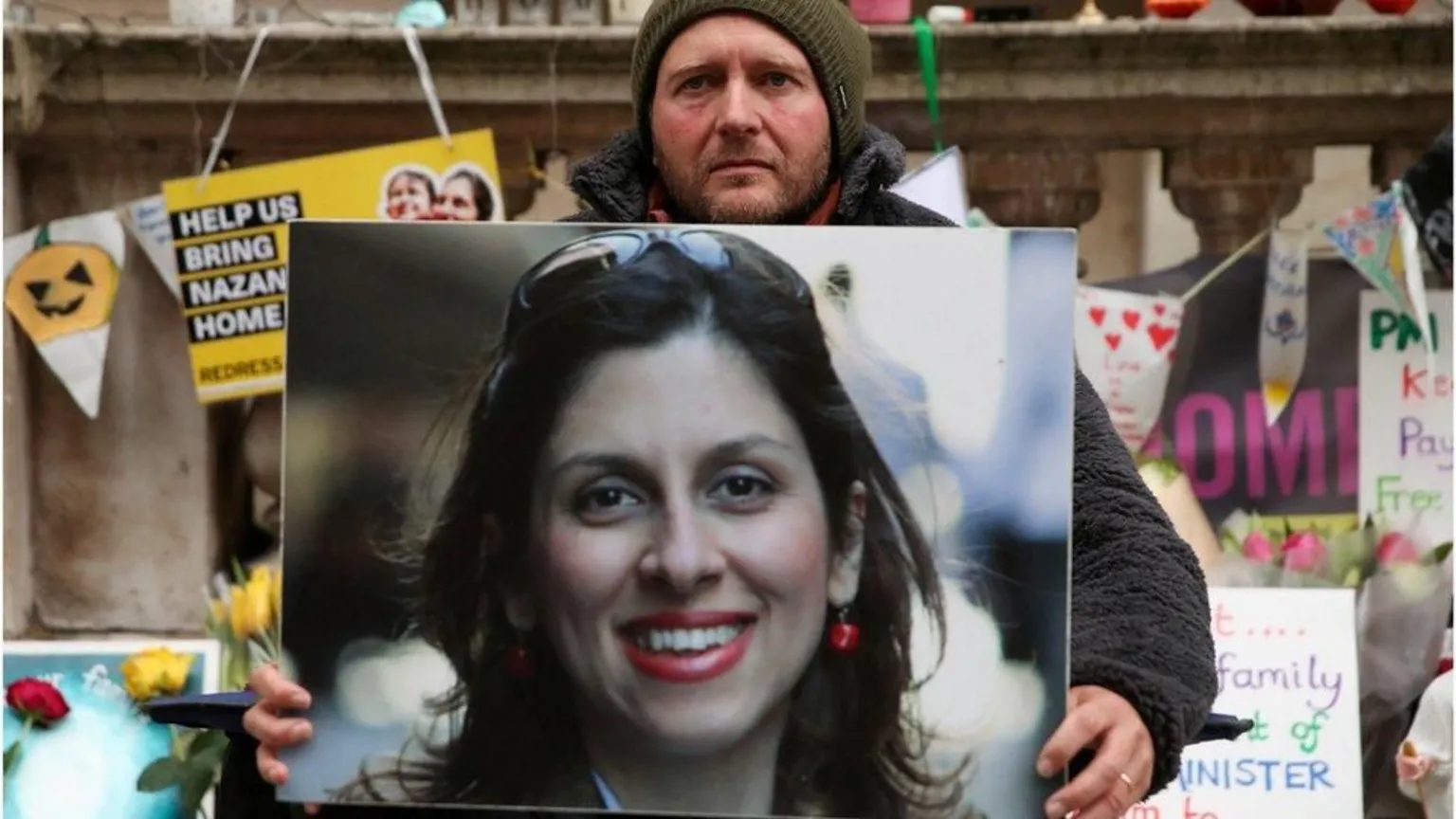  A bearded man in a beanie hat holding a massive photo of a woman with dark hair and sunglasses on her head. Behind him are various posters and flowers.