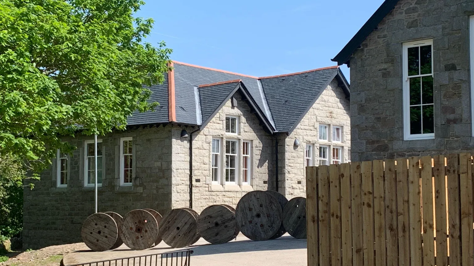 A view through trees and a wooden fence to a granite bunkhouse building. There is a row of wooden spools in front of it.