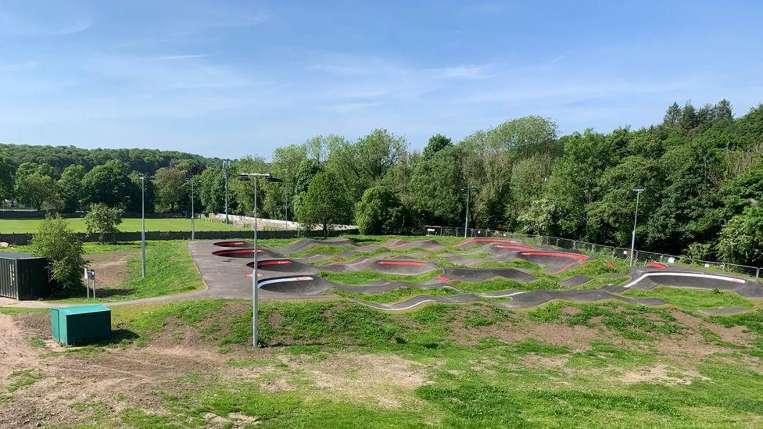A bicycle pump track surrounded by trees, lawns and a football pitch in the background