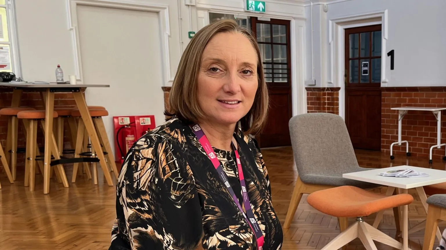 Career Connect A white woman with a brown bob haircut and wearing a patterned top and pink and purple lanyard sits on a chair centre-frame smiling at the camera. She's in a hall with other chairs and tables in the background. 