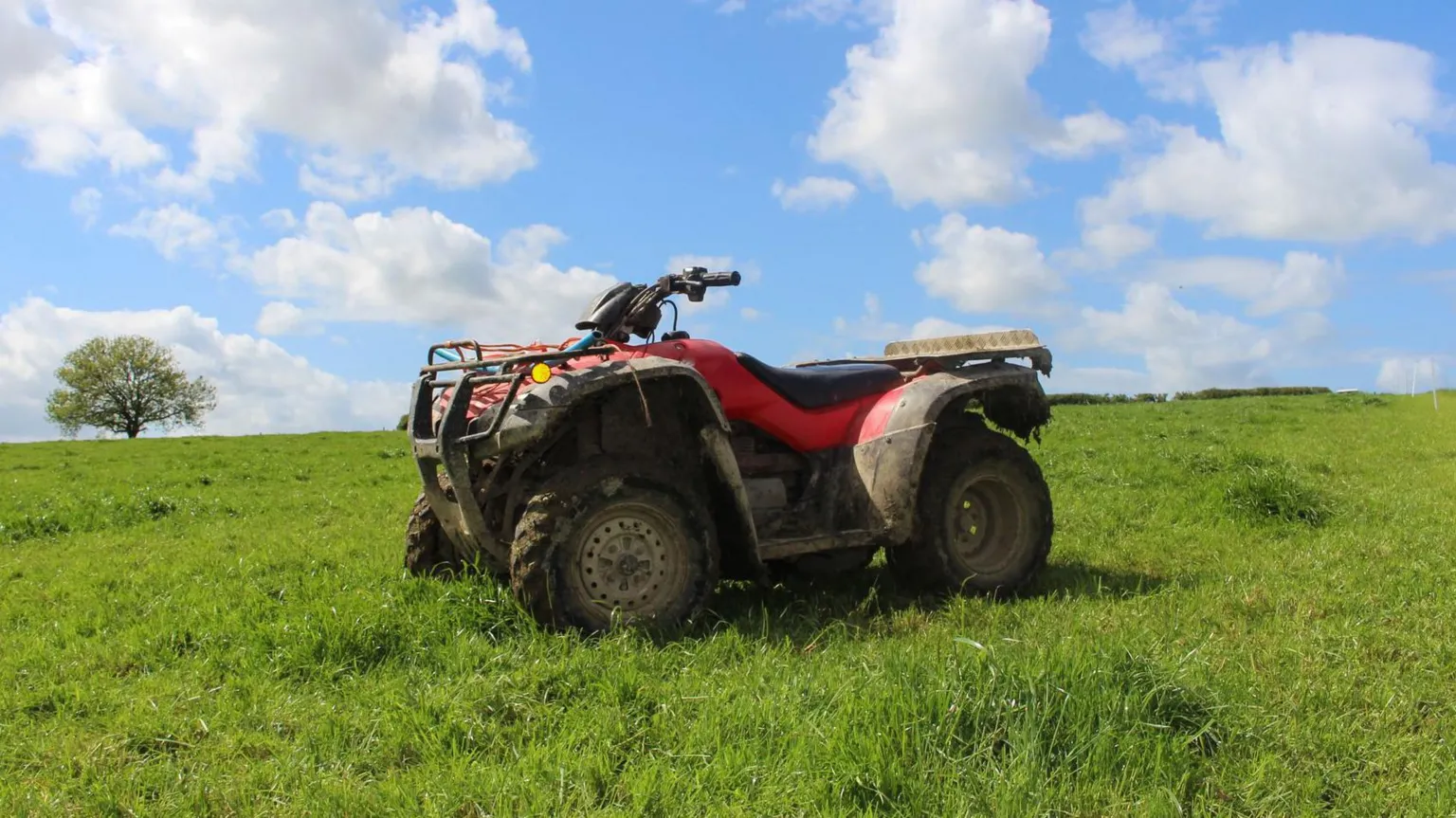  A quad bike in a field. It is a sunny day, the sky is blue with white clouds. The quad bike is in the middle of the photo, it is black and red.