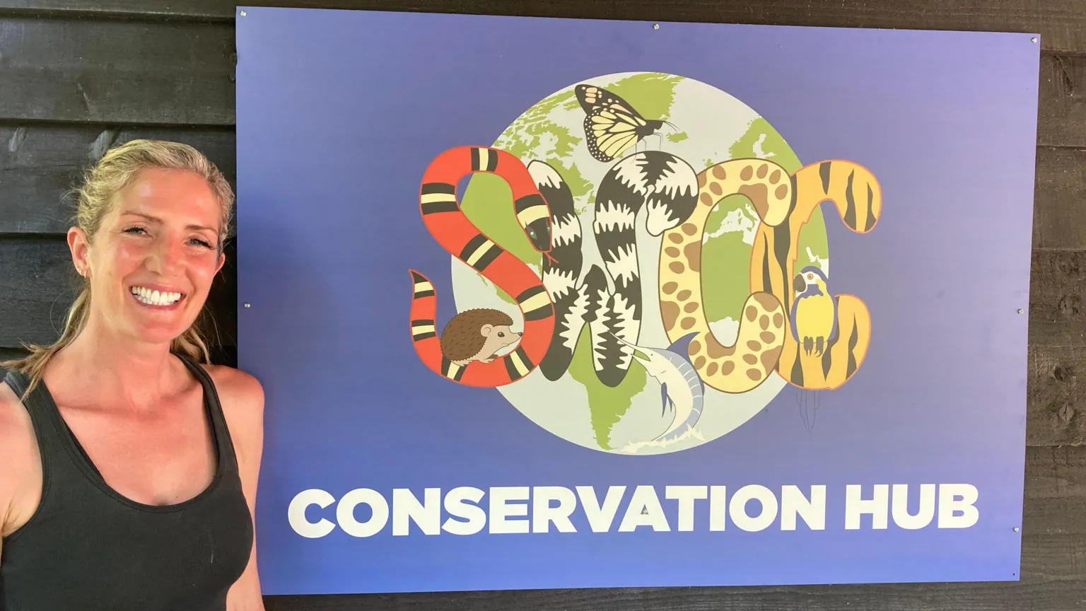 Josh Lovatt/BBC Rebecca Willers is standing in front of a wooden hut. She has long blonde hair in a ponytail and is wearing a black vest top. She is standing next to a blue sign which says CONSERVATION HUB and has several animals on it.