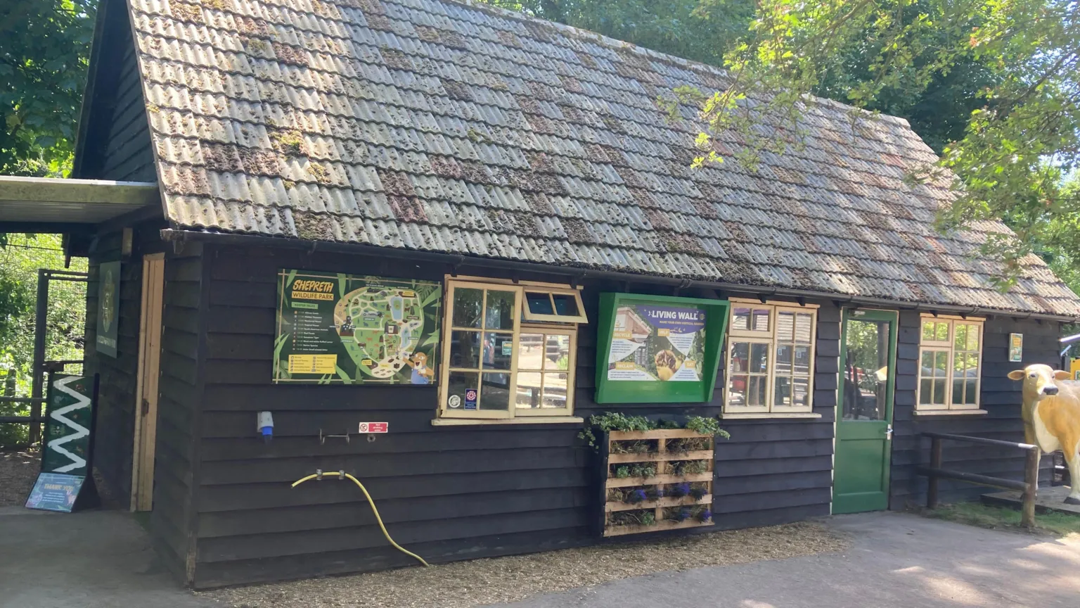 Josh Lovatt/BBC A single-storey wooden lodge is surrounded by trees and has several information boards, including a map of Shepreth Wildlife Park, on the outside walls. There is a green door to the right and there is also a statue of a cow.