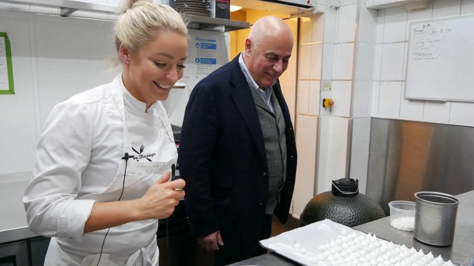 Claire Hutchings Claire Hutchings, wearing a white chef top, and with blonde hair pulled back in a bun, looks at a metal worktop, standing next to a shaven-headed man in a kitchen.