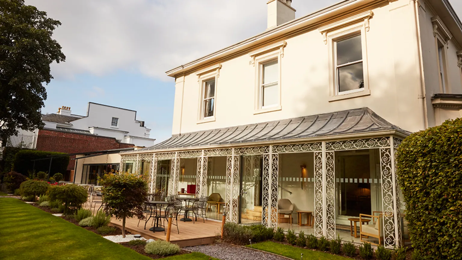 Jodi Hinds/Simpsons Cream-coloured two‑storey Georgian building with an ornate ironwork veranda overlooking a terrace with seating and lawn. Three sash windows are on the second story and ornamental trees and bushes line the garden. 