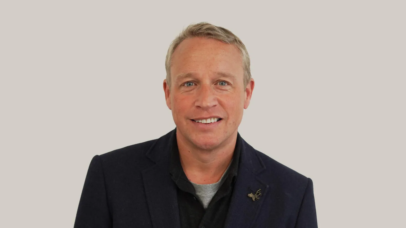 Handout A man with short hair smiles while looking at the camera. He wears a suit jacket, black overshirt and a grey t-shirt.