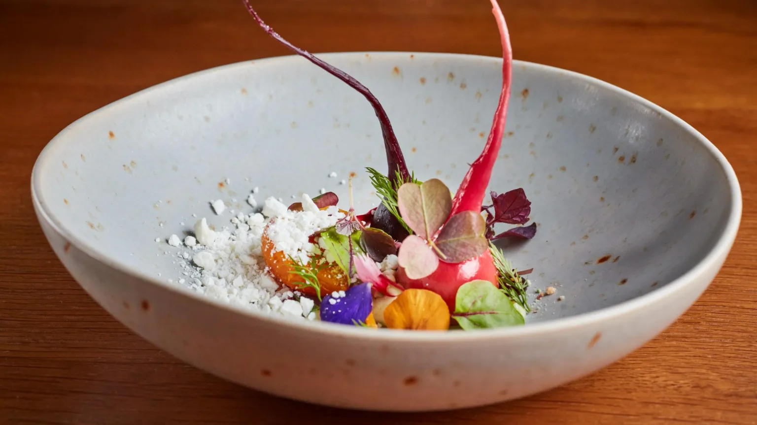 Jodi Hinds/Simpsons A small, artistic dessert with a red beet, colourful small leaves and flowers, and white crumble is served in a grey bowl on a wooden table. 