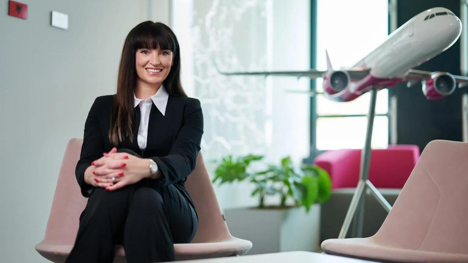 Wizz Air Yvonne smiling while wearing a suit and sitting in a pale pink chair, in front of a large model of an airplane.