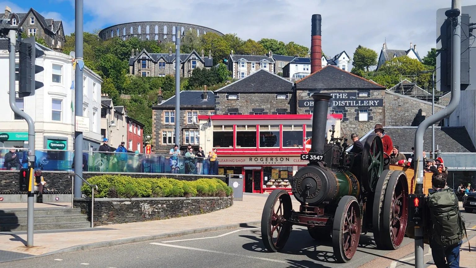 Rachel Rennie A green traction engine with red wheels is driving through a town centre, stopping at traffic lights. There is a distillery, stone-built shops, trees and a tower on a hill in the background.
