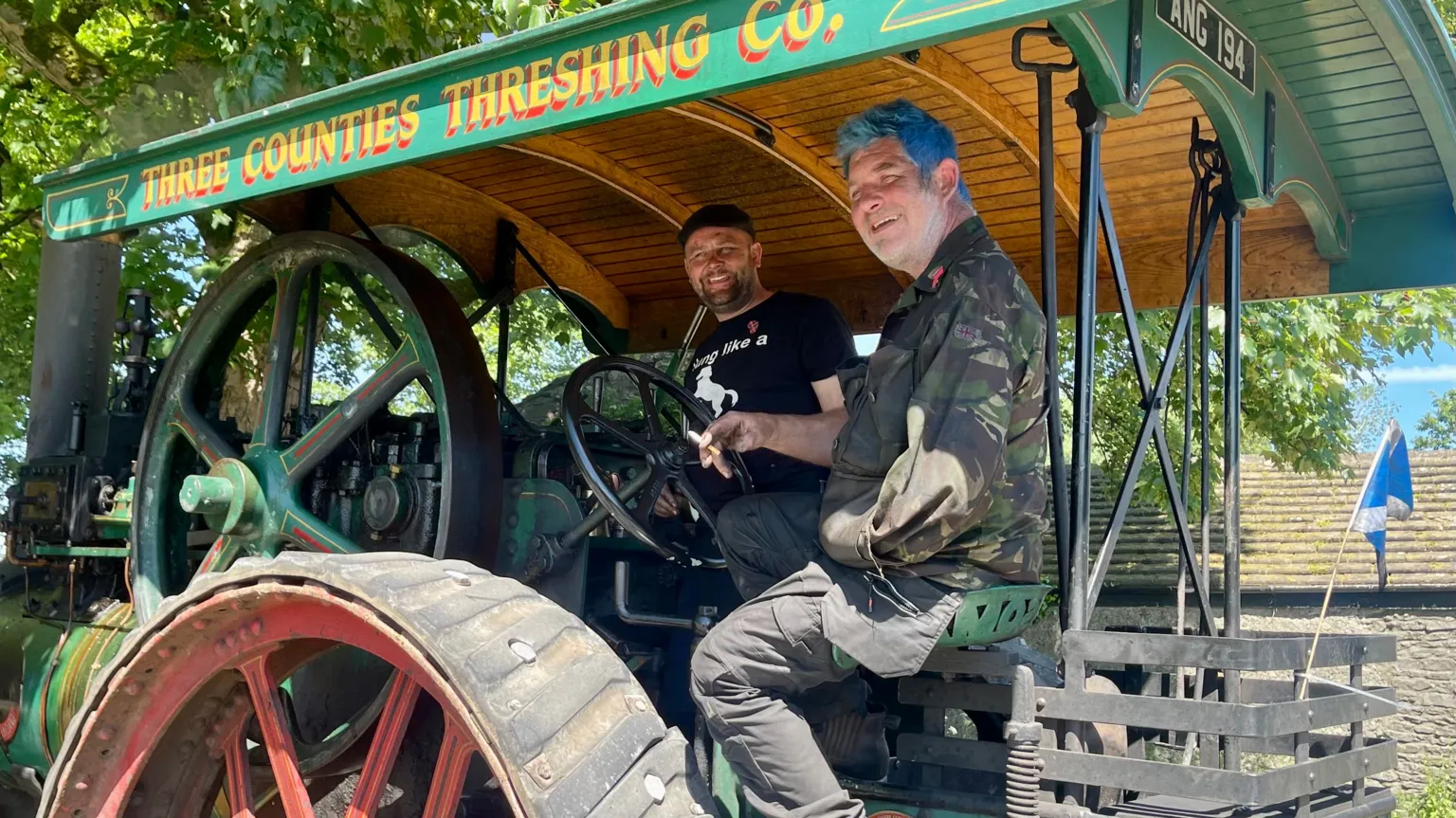 Two men sat on a green traction engine with red wheels.