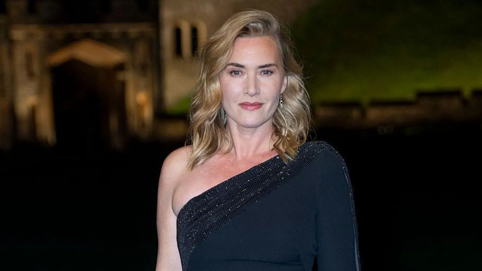  Kate smiles while looking at the camera. She is wearing a off the shoulder black sparkly dress and has blonde curly hair.