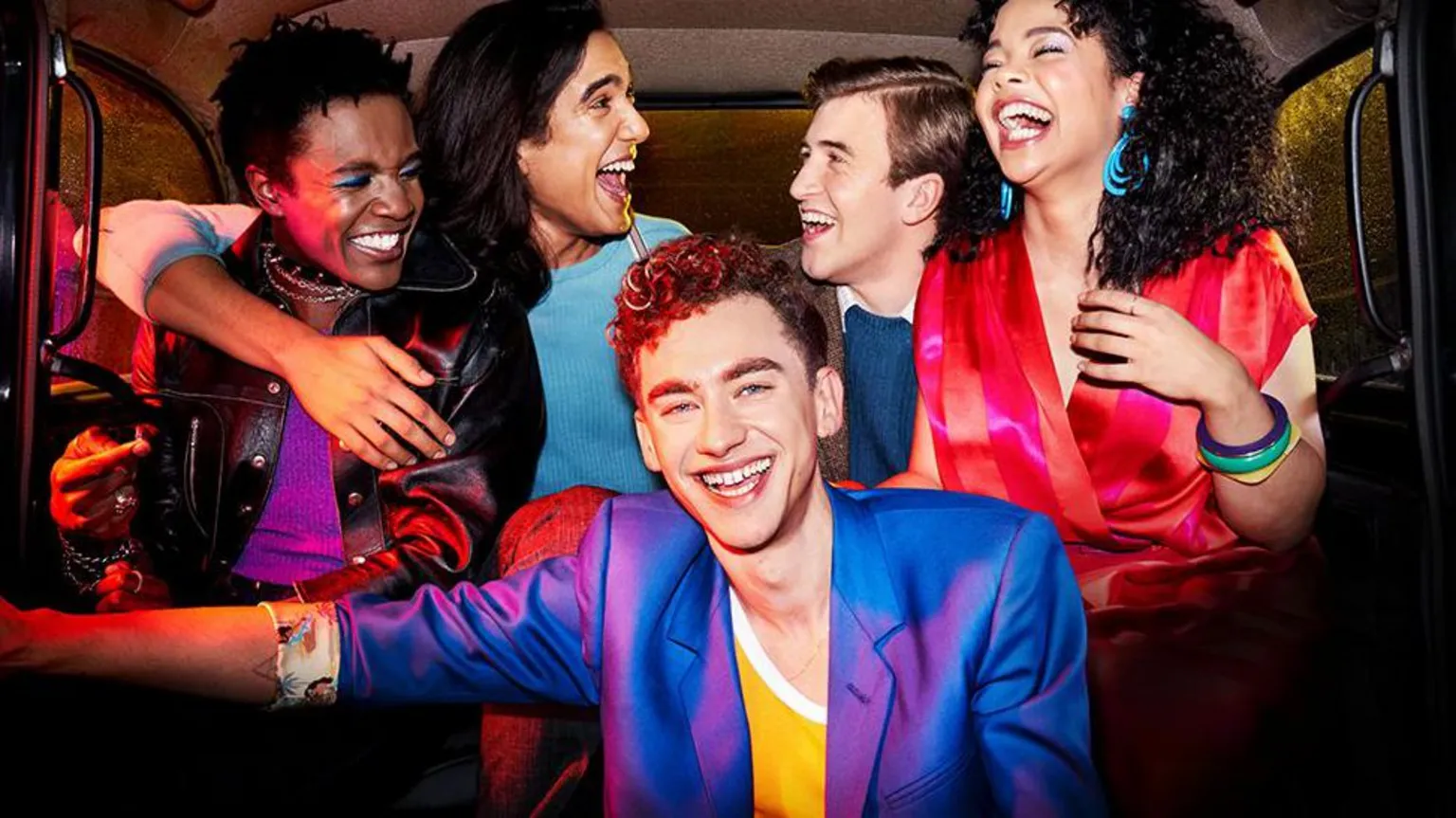  Singer and actor Olly Alexander (front) in a purple silk jacket and yellow and white t-shirt lead the cast of 's It's A Sin. He is pictured in a scene from the show in the back of a black cab with four other characters. They are all smiling.