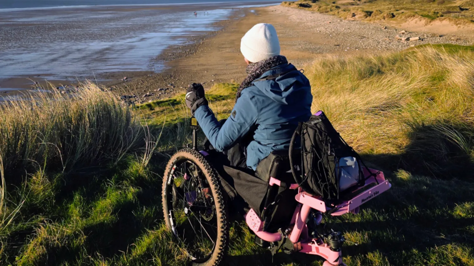 Bethany Handley Bethany in a motorised all-terrain trike wheelchair, which has a pink metal structure supporting a back wheel. She is photographed from behind wearing a blue coat, cream hat and grey scarf and gloves, looking down from the common over Sker Beach towards Swansea Bay.