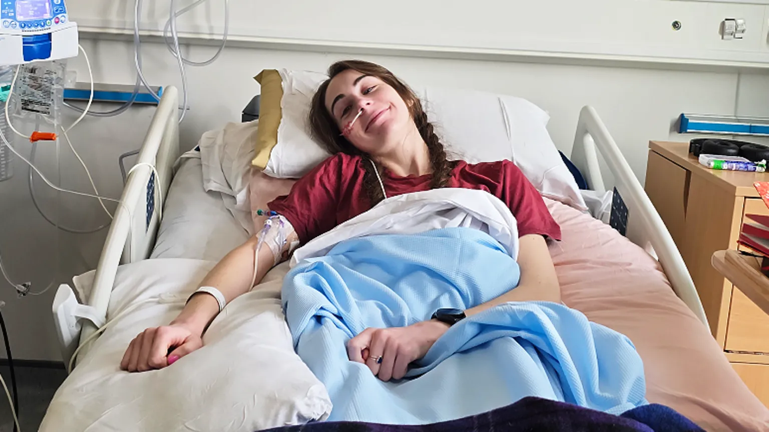 Bethany Handley Bethany lies in a hospital bed. Her long hair is plaited and she wears a dark red top. She has blankets covering her, a cuff around her upper right arm with tubes coming from it and a hospital identity bracelet on her wrist. There is a stand with medical monitors and wires to the left of the bed in the picture. To the right of the picture are a chest of drawers and a movable hospital table with a number of books on it. 