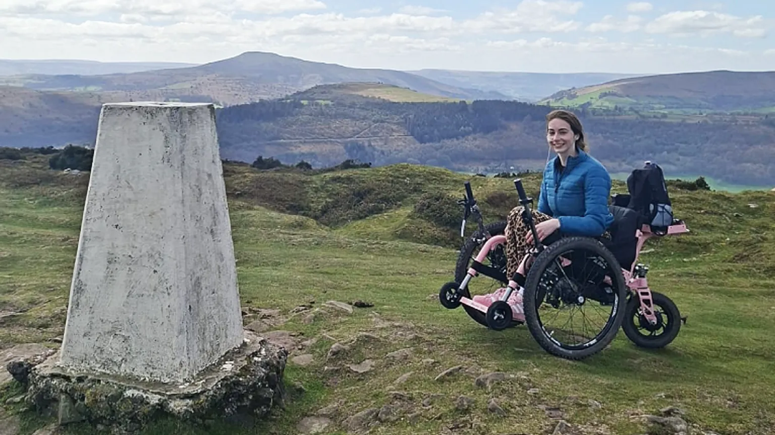 Bethany Handley Bethany in her all-terrain trike, which has pink metalwork, two large off-road wheels and a third motorised wheel at the back. She wears a blue down jacket and leggings with pink trainers. There is a trig point to the left of the trike. More hills can be seen in the background.
