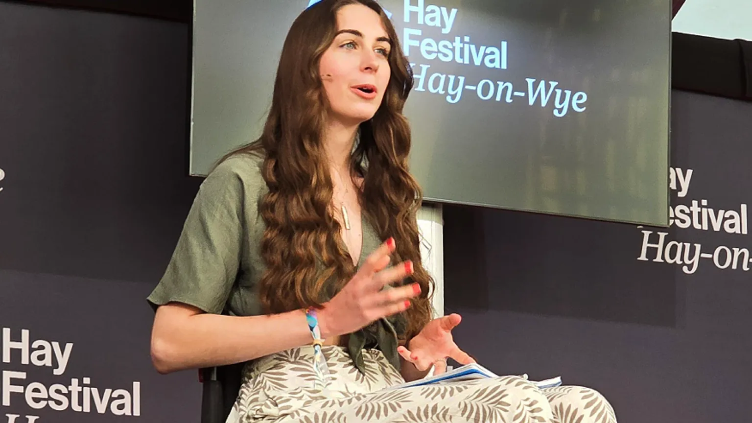 Bethany Handley Bethany on stage at Hay Festival. She wears a cream dress with a leaf pattern on it and a khaki short sleeved tie top. There is Hay Festival branding on the stage behind her. She is mid-speech looking at the audience to the right of the photo.