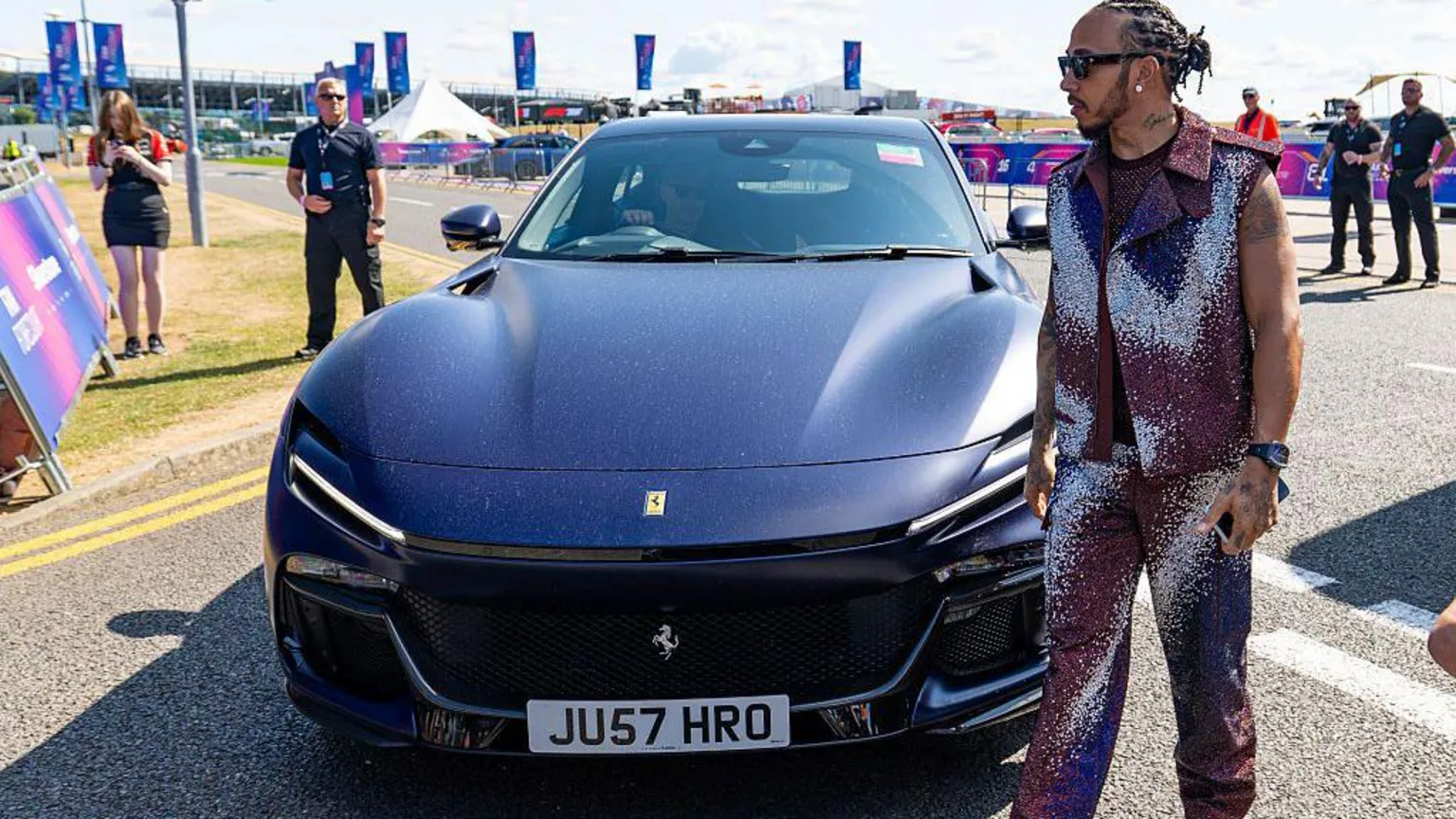  Dressed in a glittery purple outfit, F1 racer Lewis Hamilton walks past a Ferrari Purosangue SUV parked on a road.