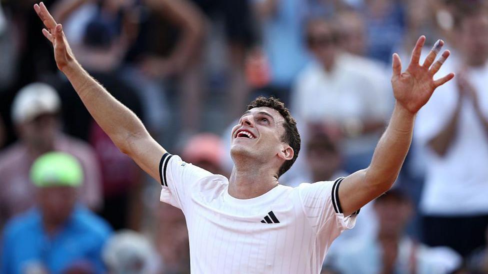 Juan Manuel Cerundolo, wearing a white t-shirt, raises his arms in the air and looks to the sky, smiling, in celebration