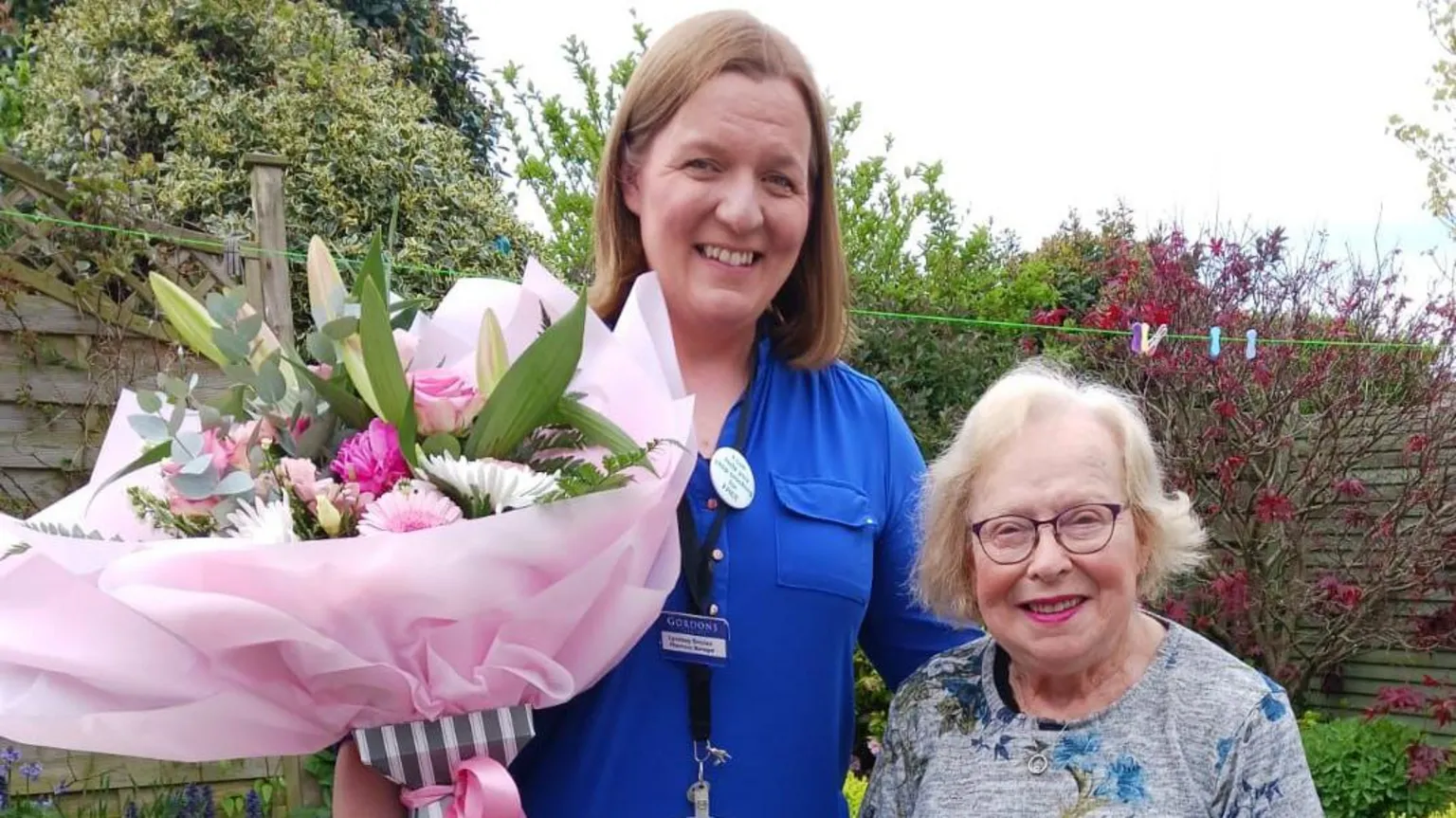 Gordons Lyndsay Sinclair has long brown hair and is wearing a blue uniform and name badge. She is holding a large bouquet of flowers in her right hand and has her other arm around Sadie. Sadie is wearing a multi-coloured top and black-rimmed glasses. 