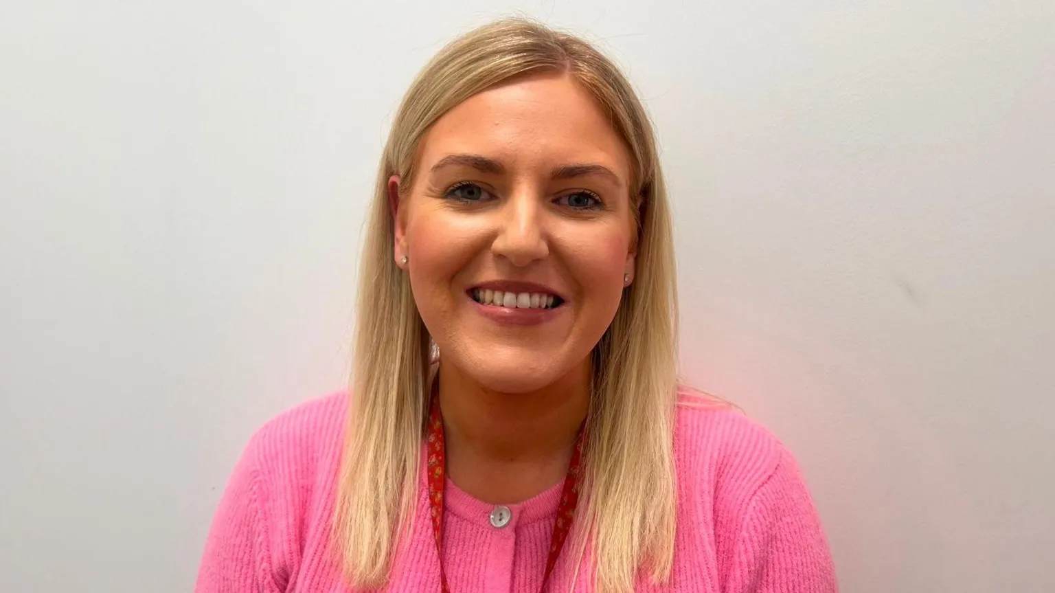 Kathryn Dobbin has long blonde hair and is wearing a pink cardigan. She is standing in front of a plain white wall. 