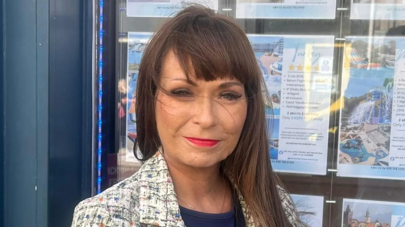 Jenny Elliott has long brown hair and is wearing a white multi-coloured jacket. She is standing in front of her travel agency shop. 