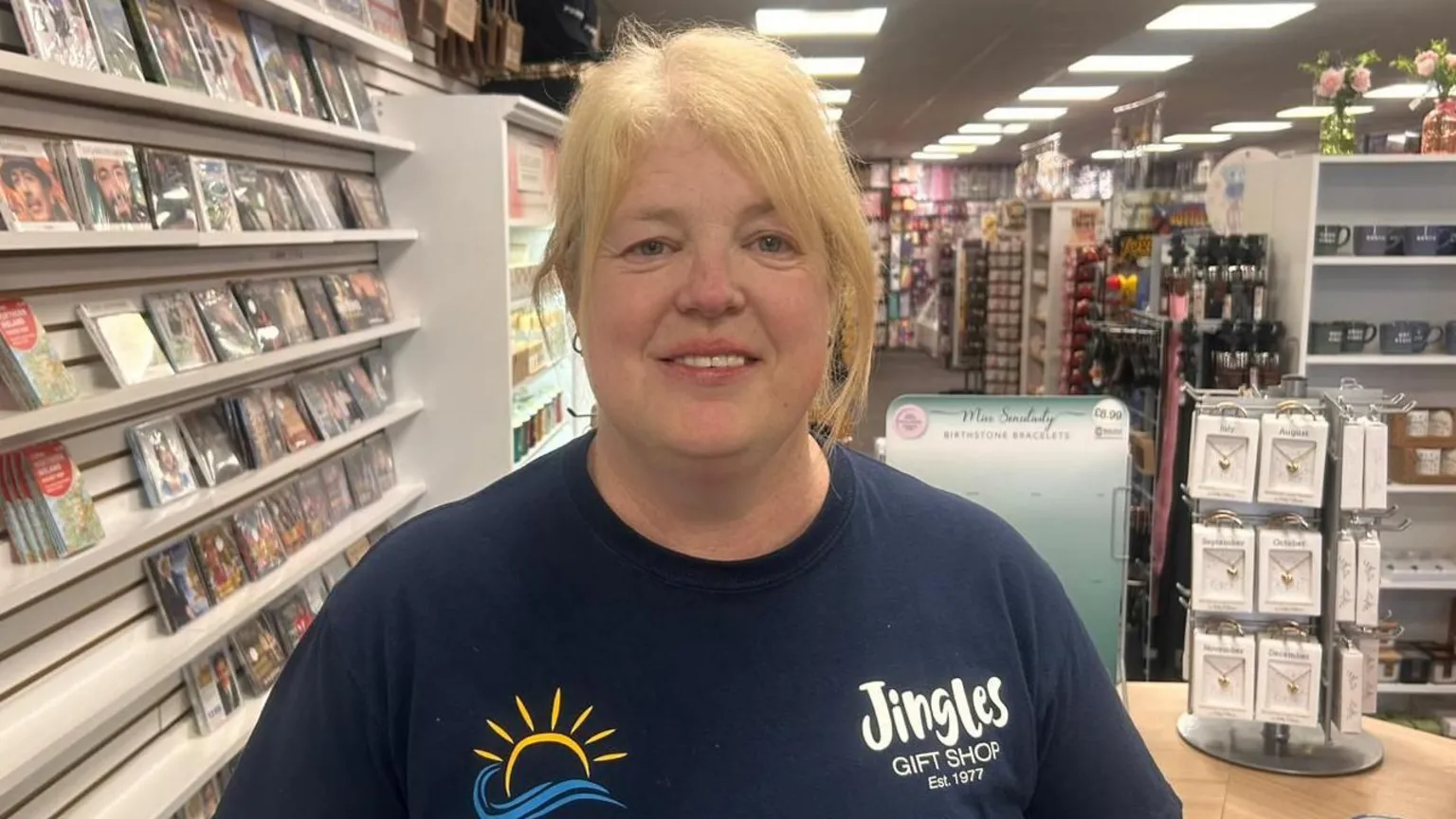 Karen McMath has long blonde hair. She is wearing a navy T-shirt with the logo Jingles and a sun logo. A number of pictures and items are visible in the store behind her.