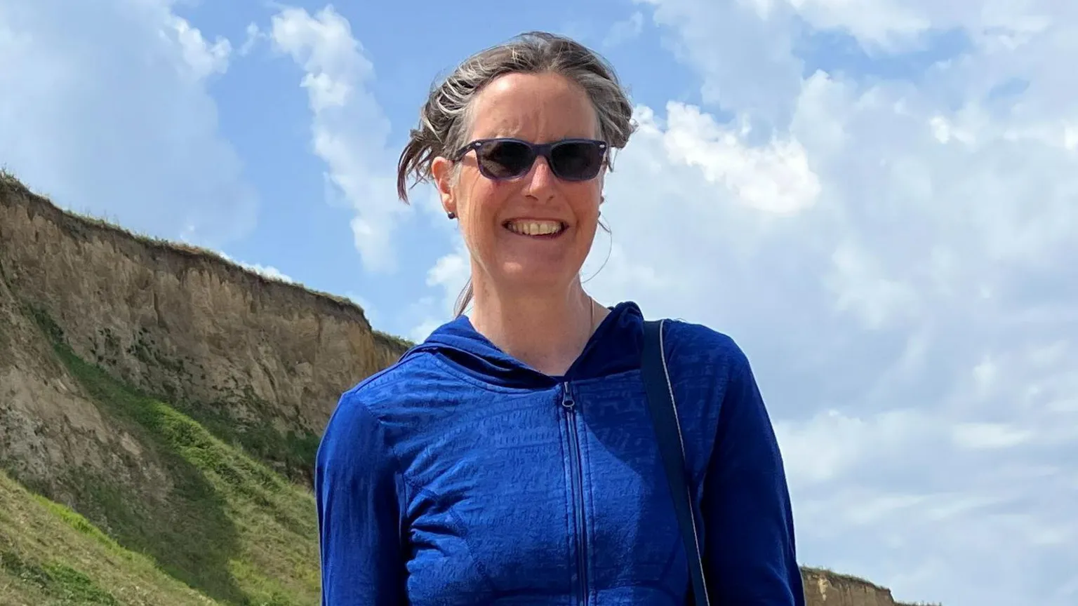 Alex Dunlop/BBC Julia James, standing on Cromer beach. She is looking directly at the camera and smiling. She is wearing a blue zipped up hoodie and a pair of sunglasses over her eyes.