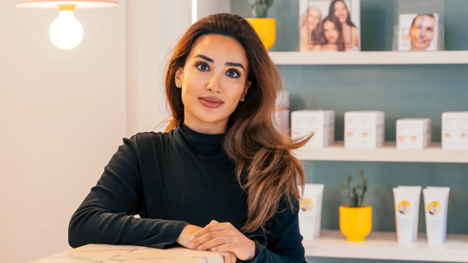 Shireen Forster A person stands behind a marble counter in a modern shop, with shelves of skincare products and colourful signs on the wall behind them. 