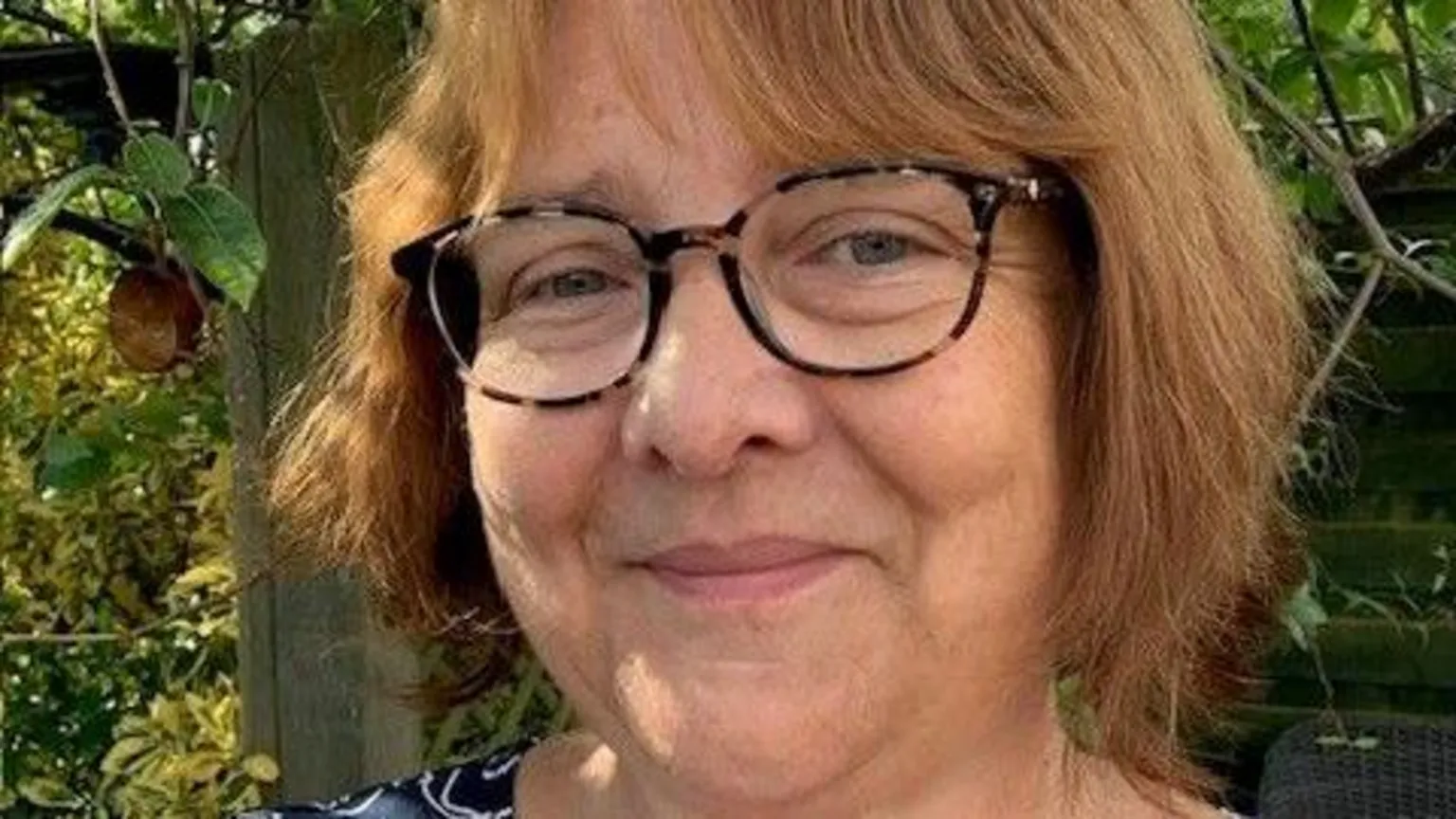 Kate Morris A woman wearing glasses poses outdoors in a garden, with green leaves and a wooden fence in the background.