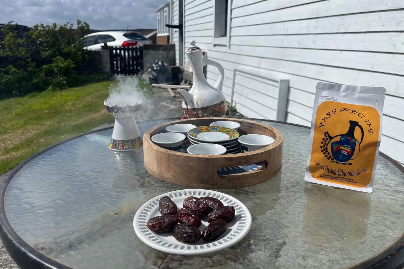 A traditional Ethiopian coffee ceremony set up on a table outside. There is a coffee pot, a bag of coffee, a plate of dates, and a smoking incense burner.