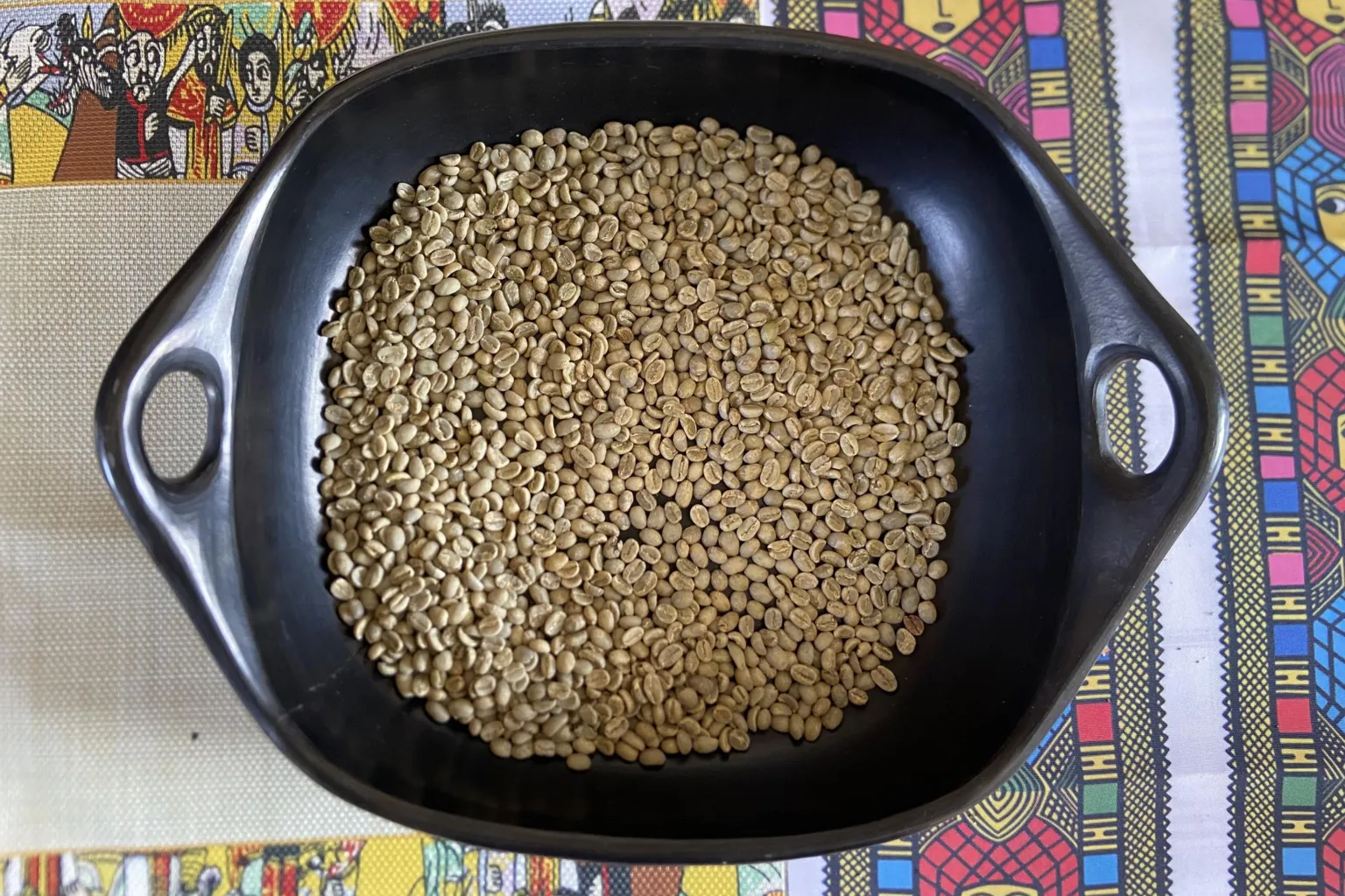 A black pan filled with pale green coffee bean sitting on top of a colourful table cloth.
