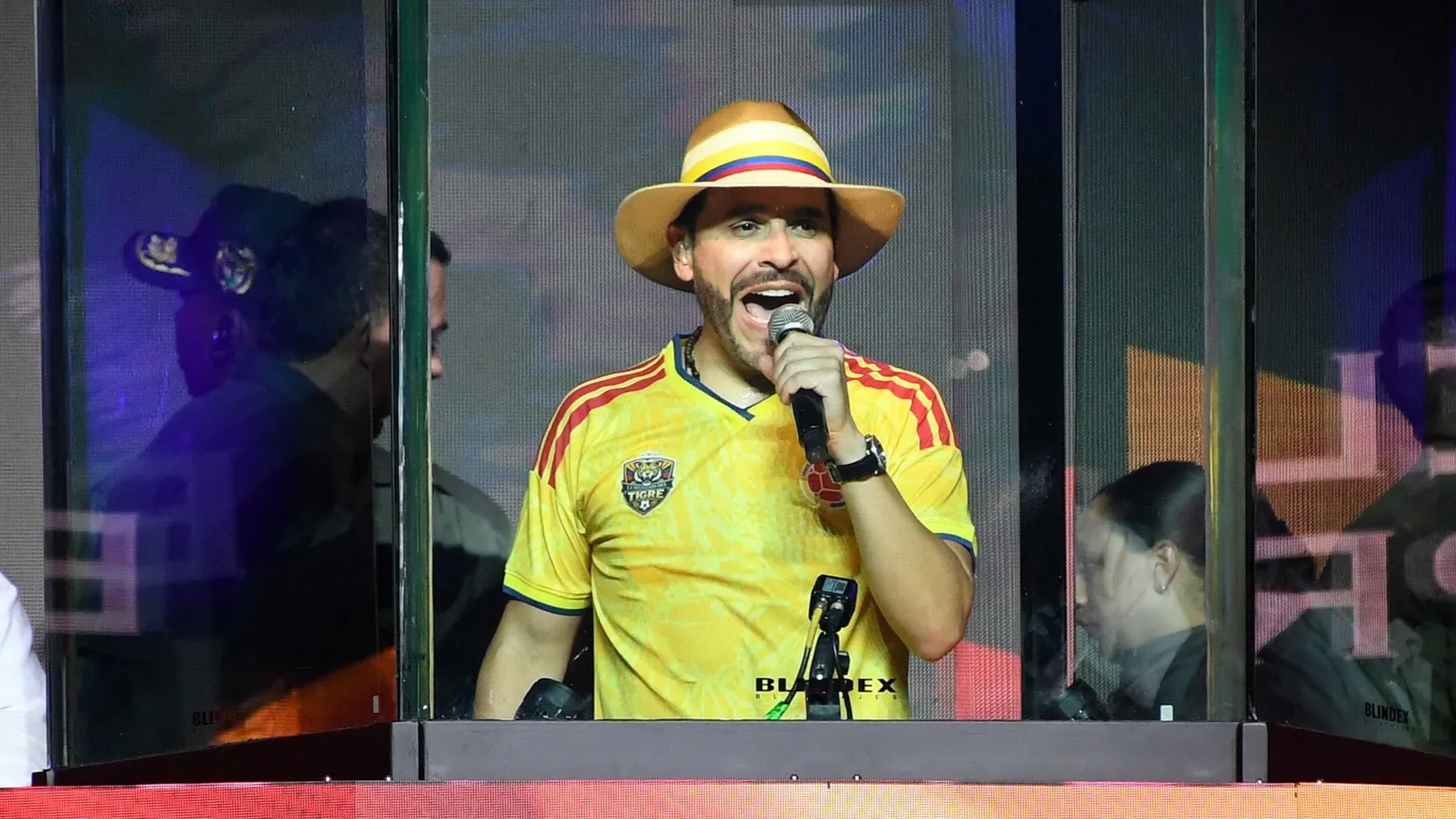  Abelardo de la Espriella speaks behind bulletproof glass during a campaign rally in Medellin. He wears a Colombia football shirt and striped hat.