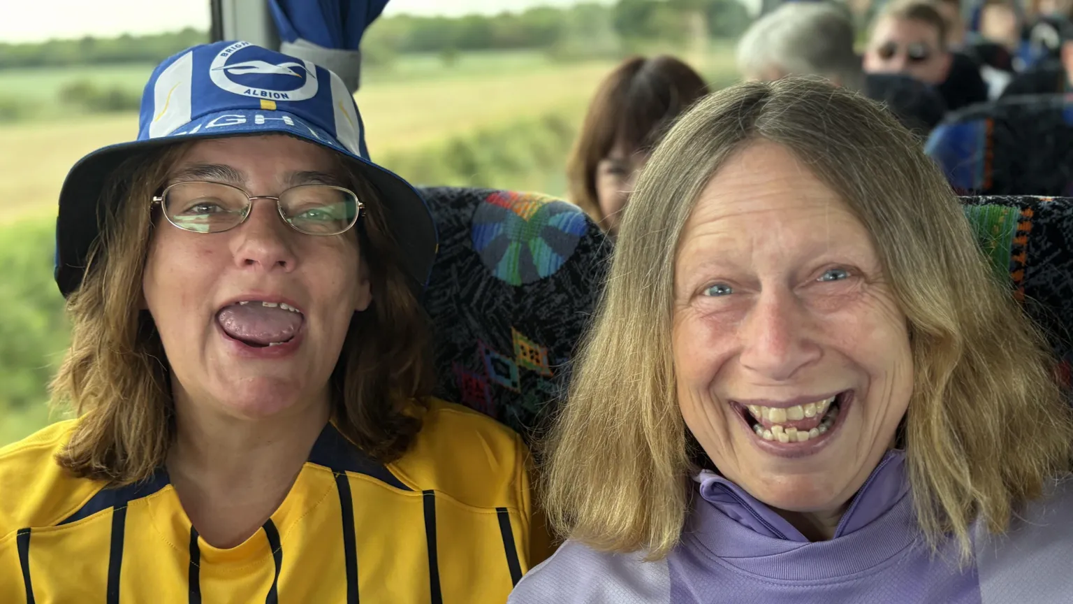 Janet Cotton and Janette Back sat on coach seats. Janet is wearing a yellow football top and a Brighton blue bucket hat. Janette is wearing a purple Brighton kit.