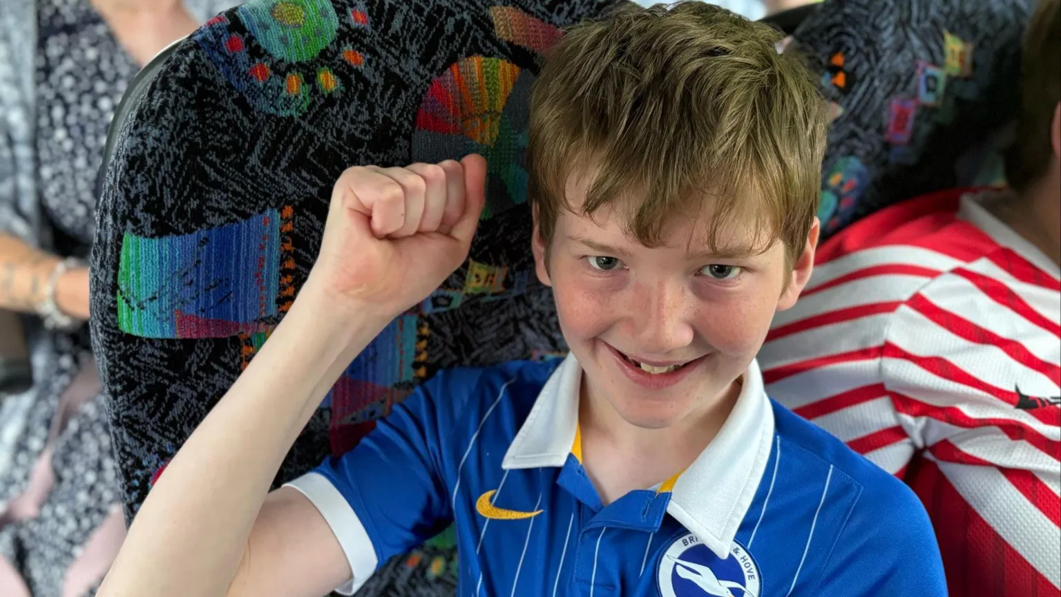 William sat on a bus seat wearing a blue Brighton football shirt. He has one arm raised with a clenched fist and is smiling.