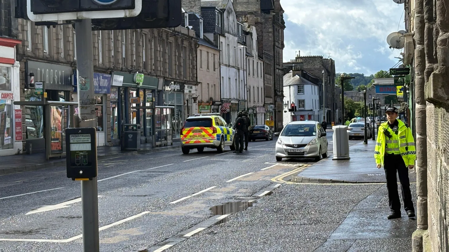 Armed police beside a police car in the middle of a street. There is a police officer in a hi-vis jacket in the foreground looking at the camera 